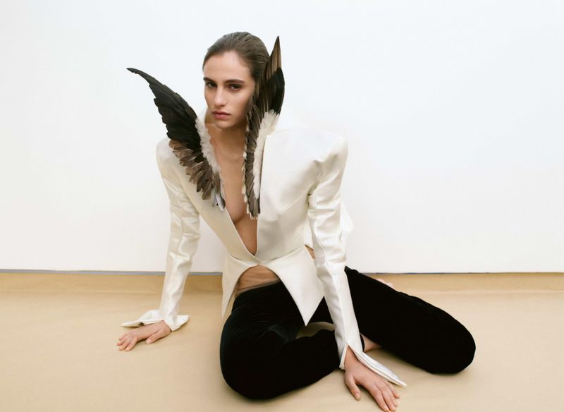 A woman with light skin poses sitting on the floor, wearing a white, long-sleeved, plunging top with black pants and feathered embellishments.
