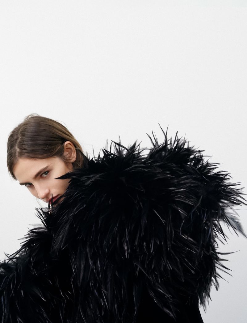 Close-up of a woman with long brown hair wearing a black feathered garment against a bright white background in a studio setting.