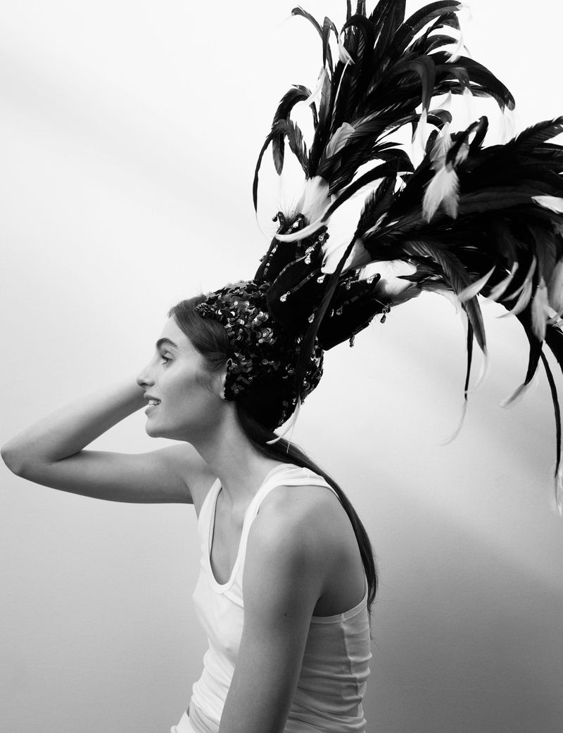 Black and white portrait of a young woman in a white tank top with a feather headdress against a neutral backdrop.