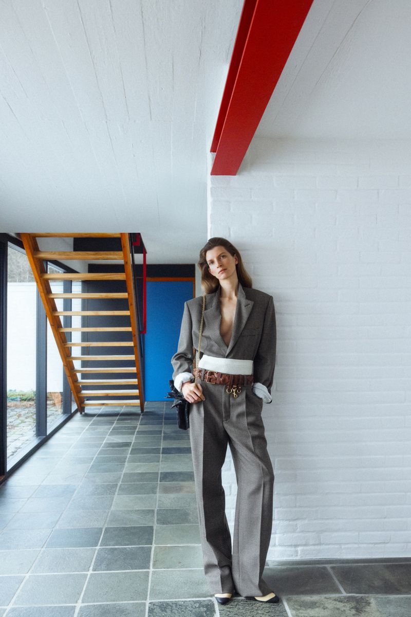 Fashion editorial shot: Woman in grey suit poses indoors with white walls, dark floors, wooden staircase, and red architectural beam.