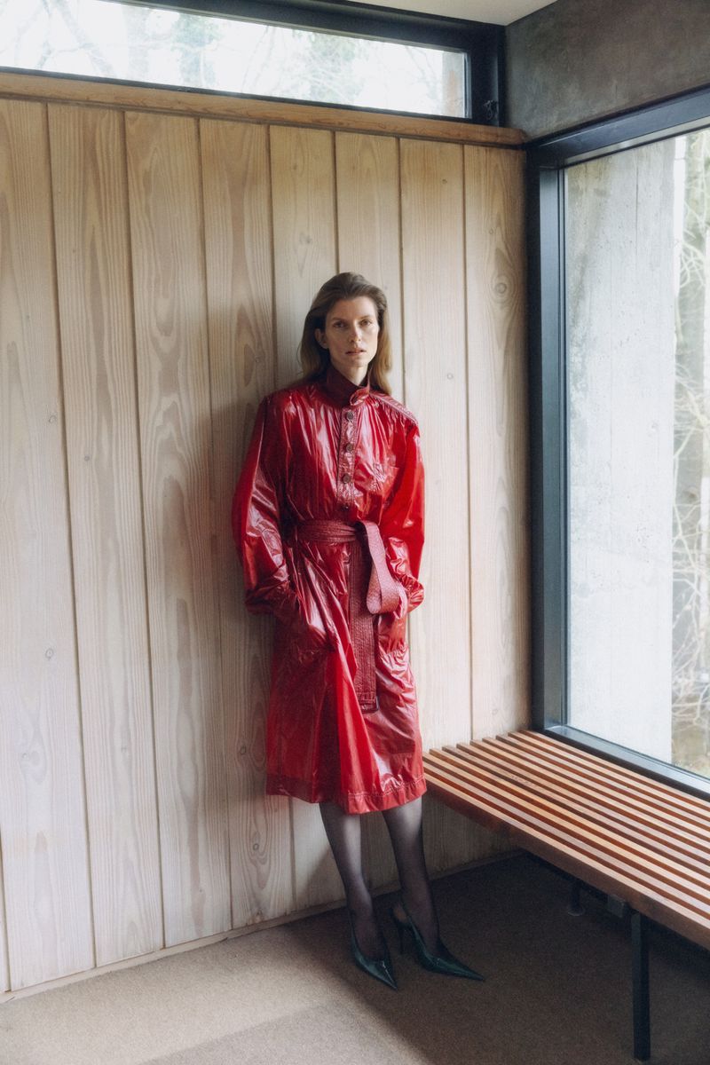 A woman with brown hair poses indoors wearing a red, belted vinyl coat, sheer tights, and black heels, standing against a wood-paneled wall.