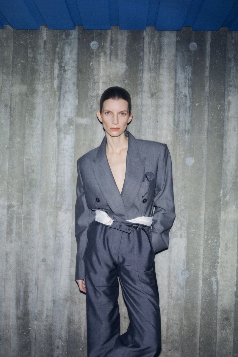 Fashion portrait of a woman in a grey pantsuit with a white belt against a textured concrete wall and blue corrugated metal ceiling.