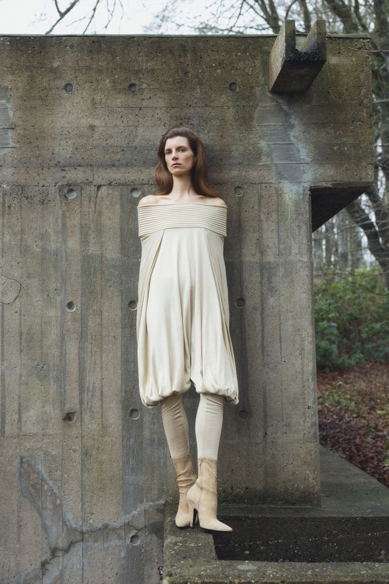 A woman in an off-the-shoulder dress stands against a concrete wall, complemented by leggings and heeled boots in an outdoor setting.