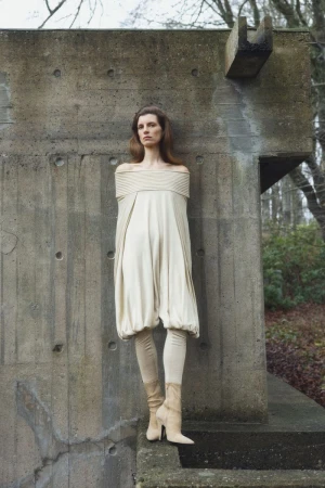 A woman in an off-the-shoulder dress stands against a concrete wall, complemented by leggings and heeled boots in an outdoor setting.