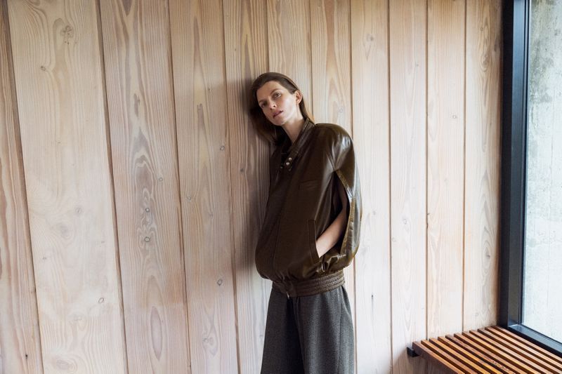 Woman in a green bomber jacket leaning against a light wood wall, wearing grey pants, indoor lighting, and a neutral facial expression.