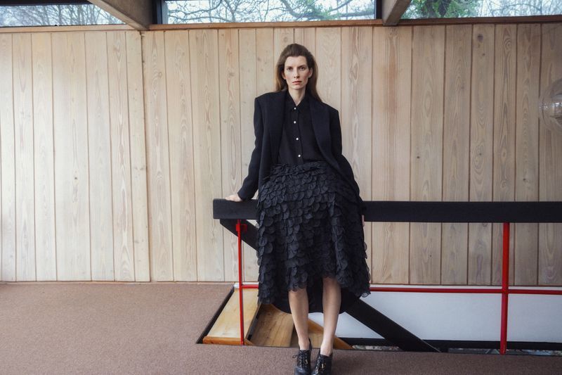 Fashion shot of a woman in a black suit and skirt posing indoors with natural lighting and a wood paneled wall in the background.