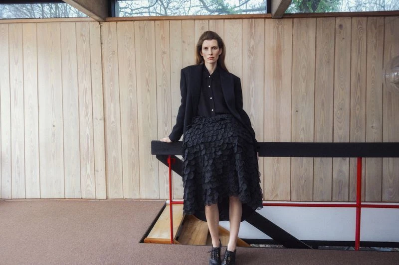 Fashion shot of a woman in a black suit and skirt posing indoors with natural lighting and a wood paneled wall in the background.