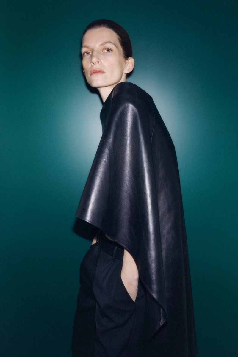 Fashionable woman in a black leather poncho poses against a teal backdrop, showcasing a modern and sleek aesthetic.