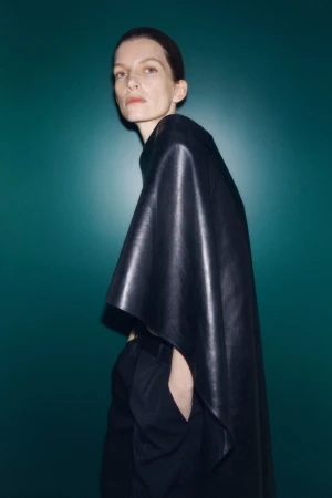 Fashionable woman in a black leather poncho poses against a teal backdrop, showcasing a modern and sleek aesthetic.