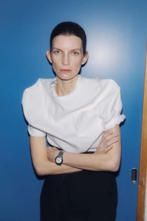 Model with dark hair wearing a structured white t-shirt and dark trousers against a blue wall