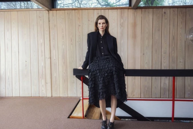 Model standing by a wooden wall wearing a black blazer, dark shirt, and a textured black skirt for Elle Denmark editorial.