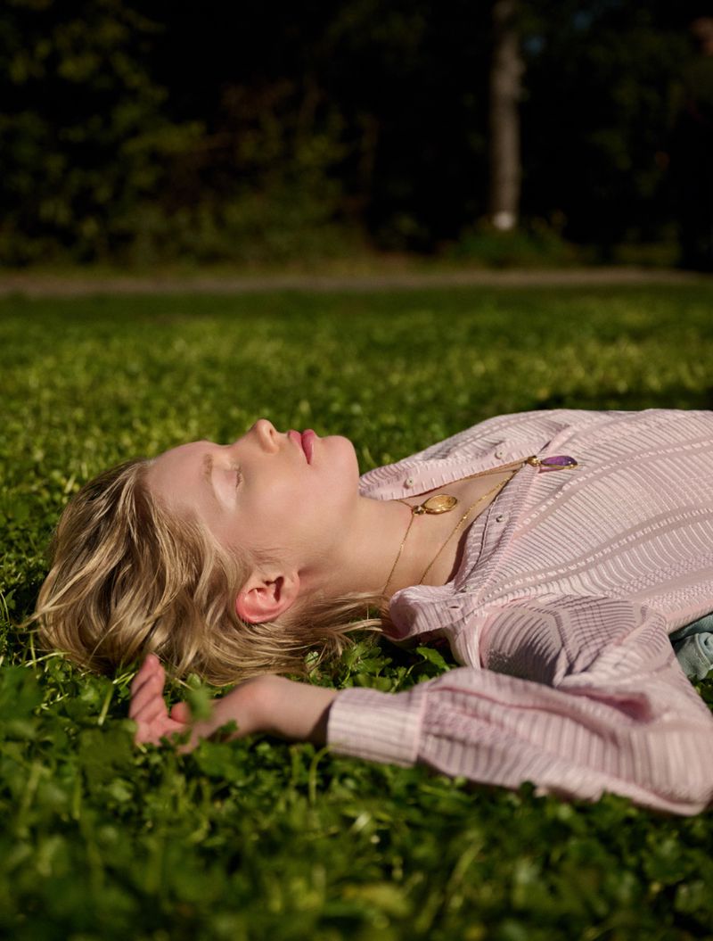 Fair-skinned woman with blonde hair relaxes in a sunny grassy field, wearing a striped pink shirt and layered gold necklaces, eyes closed.