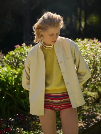 Blonde woman poses outdoors wearing yellow top, tan jacket, and striped shorts, with green foliage in the blurred background.