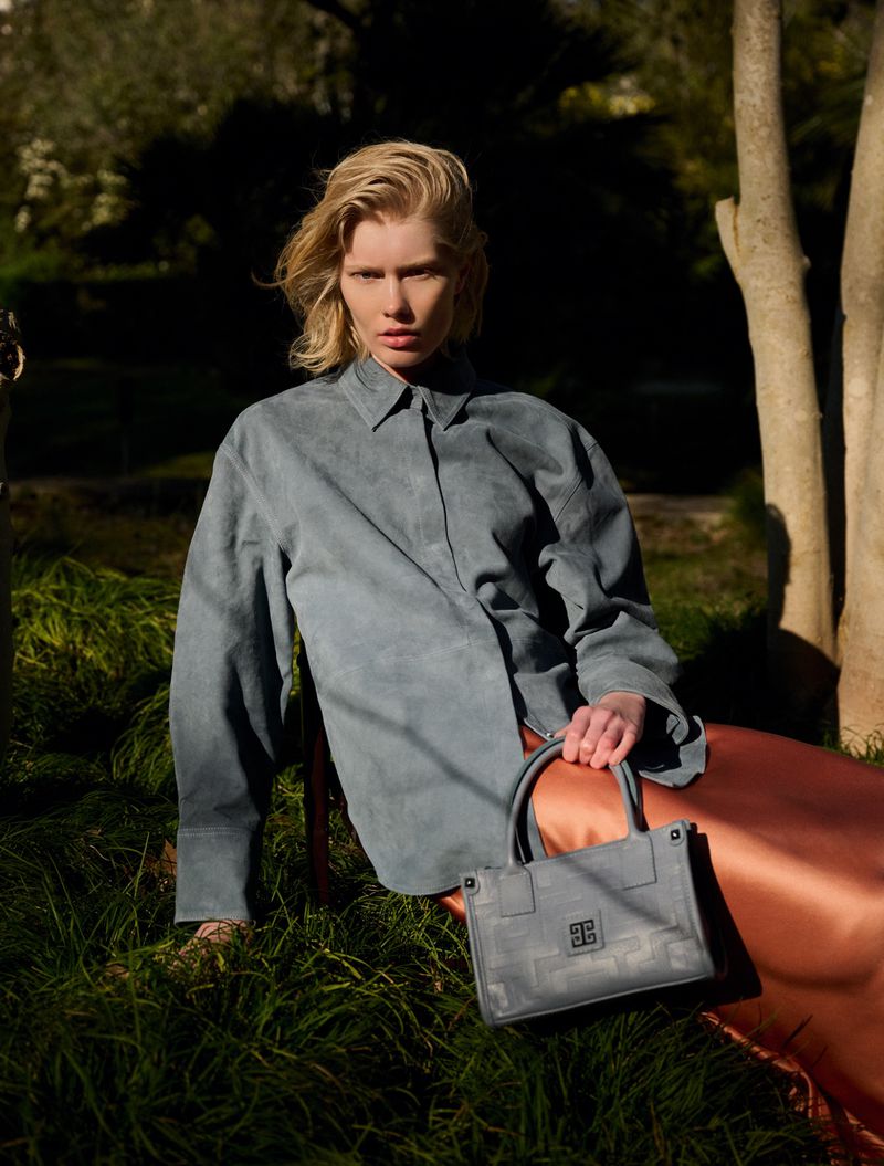 Elegant portrait of a blonde-haired woman outdoors wearing a blue button-up shirt and holding a structured handbag, in a natural setting.