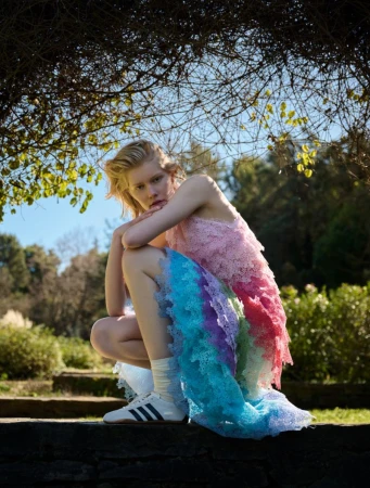 Woman with blonde hair poses outdoors wearing a colorful, tiered gown and sneakers, sitting on a stone structure under a tree canopy.