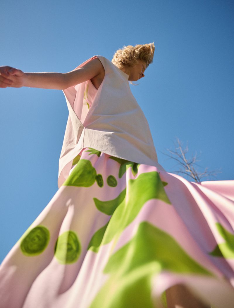 Woman in floral skirt stands against blue sky, arms outstretched, in bright sunshine. Fashion shot, low angle from below.