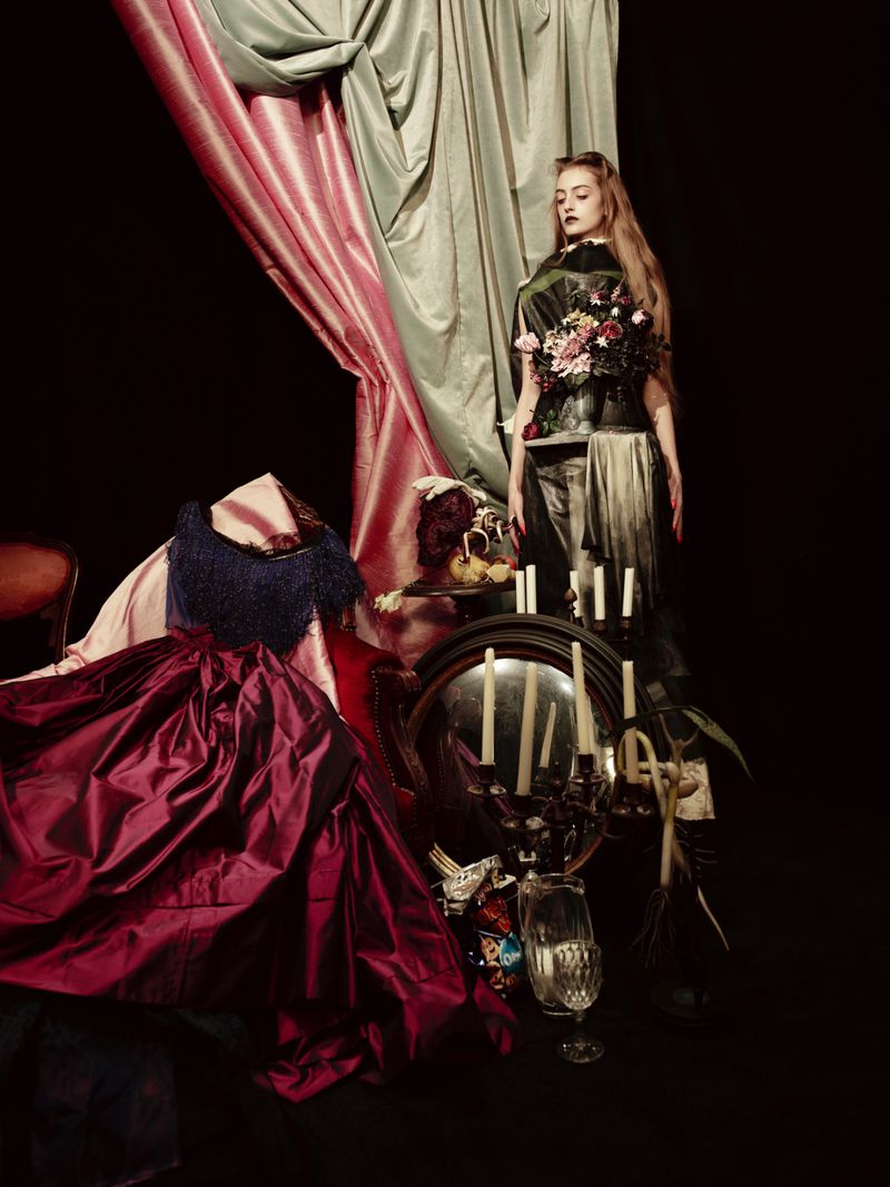 Woman in ornate costume stands in a dark photo studio amid draping fabrics, candles, and antique objects, creating an editorial fashion portrait.