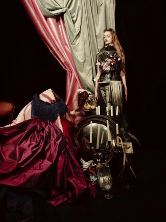 Woman in ornate costume stands in a dark photo studio amid draping fabrics, candles, and antique objects, creating an editorial fashion portrait.