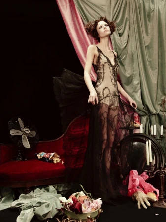 A slender woman poses in an embellished black lace slip dress, standing near an antique chaise lounge and ornate decorations.