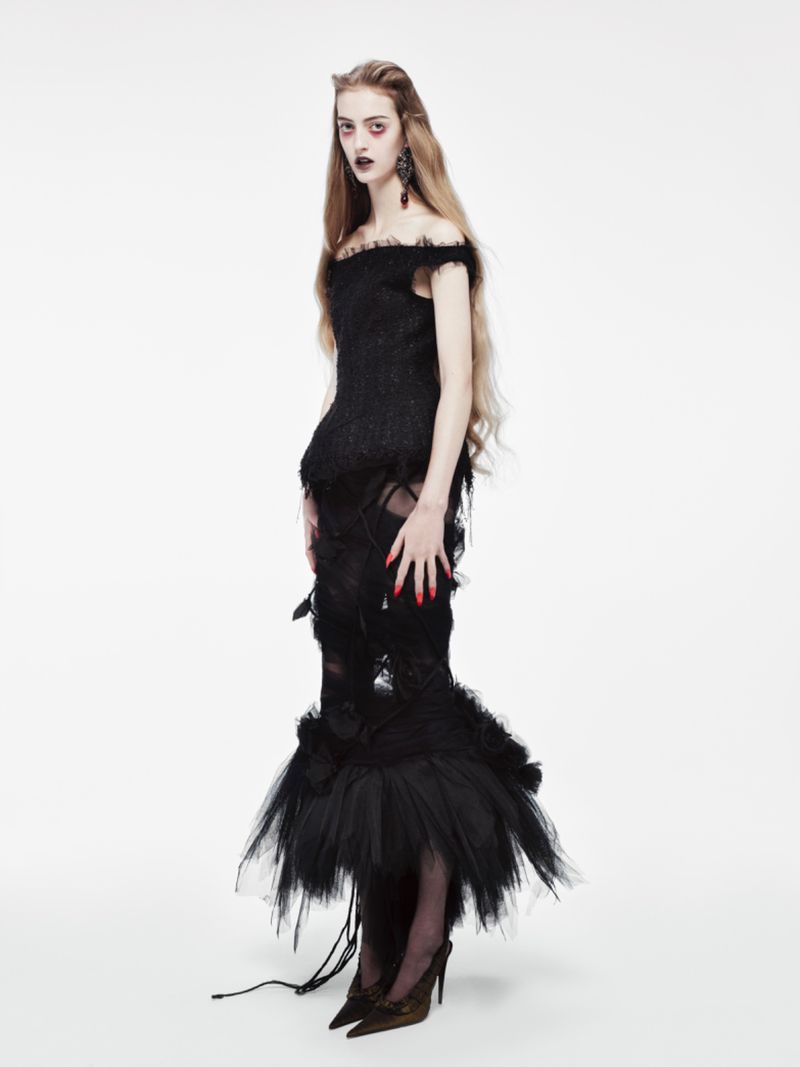 Fashion shot of a woman in a black dress with long blonde hair against a white background, showcasing dark makeup and unique attire.