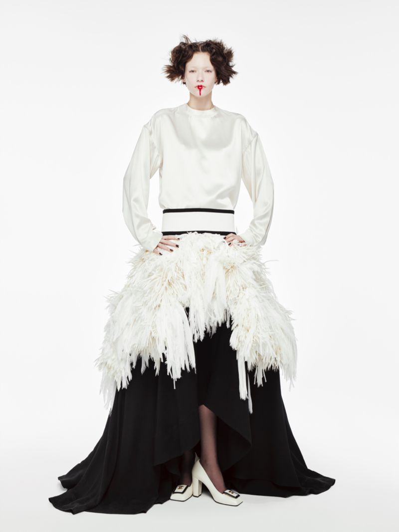 A fair-skinned model poses in a white long-sleeved top, a feathered skirt, and white heels against a white backdrop in a studio shot.
