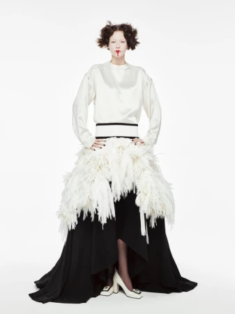 A fair-skinned model poses in a white long-sleeved top, a feathered skirt, and white heels against a white backdrop in a studio shot.