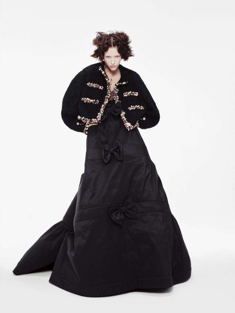 Woman in black taffeta gown and embellished jacket poses in a fashion editorial shot on a white background in a photo studio.