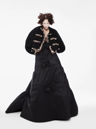 Woman in black taffeta gown and embellished jacket poses in a fashion editorial shot on a white background in a photo studio.