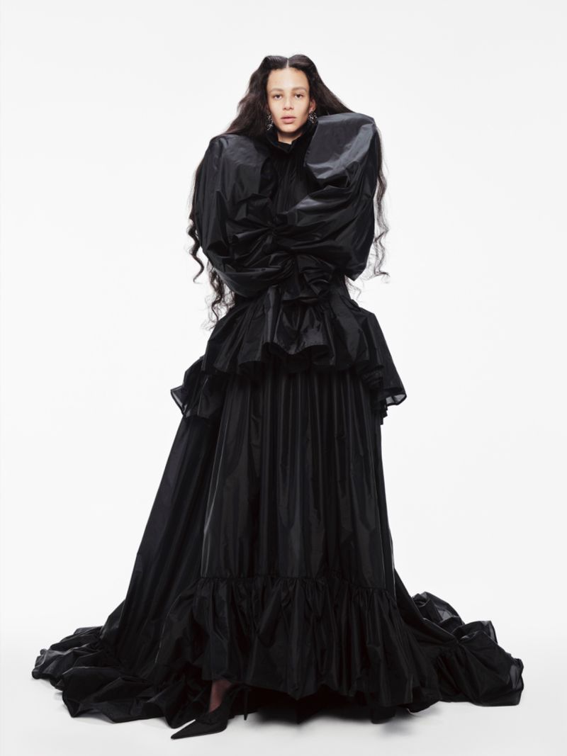 Young woman wearing an elaborate long, black ruffle gown with puffy sleeves in a studio setting against a white background.