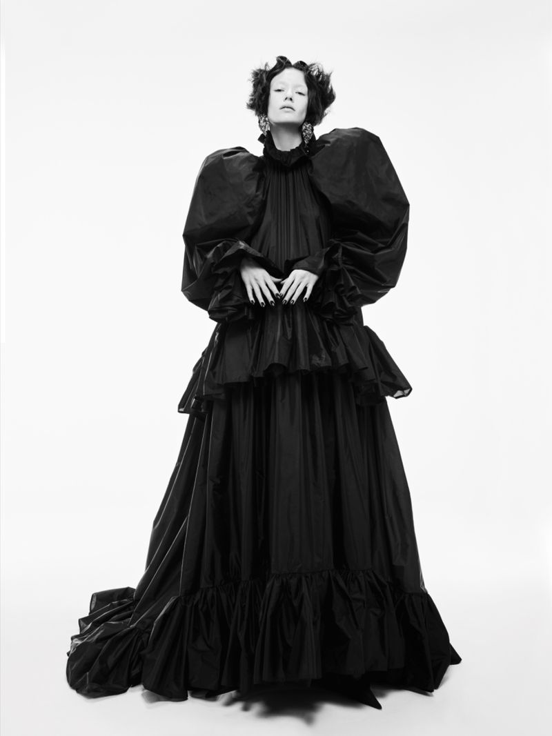 A fair-skinned person in a black Victorian gown with puffy sleeves poses against a bright white backdrop, full studio shot.