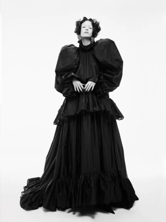 A fair-skinned person in a black Victorian gown with puffy sleeves poses against a bright white backdrop, full studio shot.