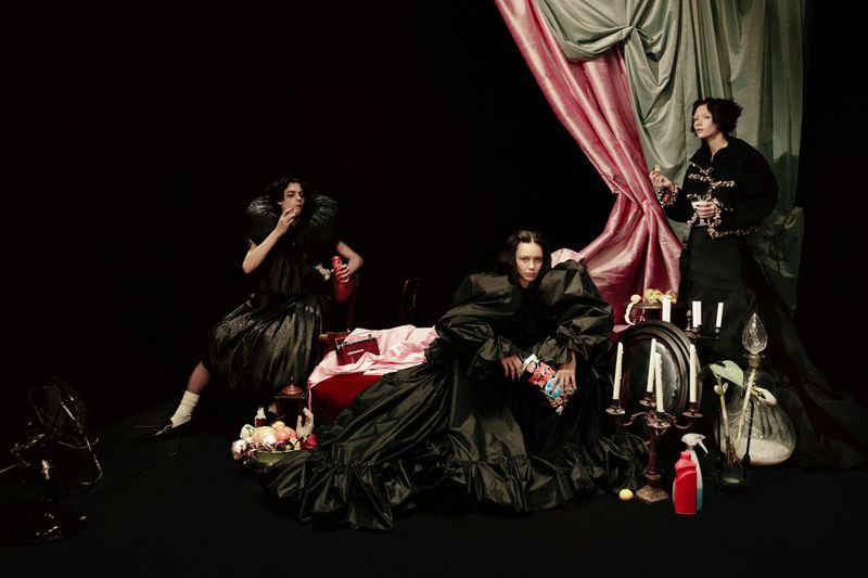 Three women in black couture gowns pose among vintage props and pink and grey draping against a dark background in a theatrical tableau.