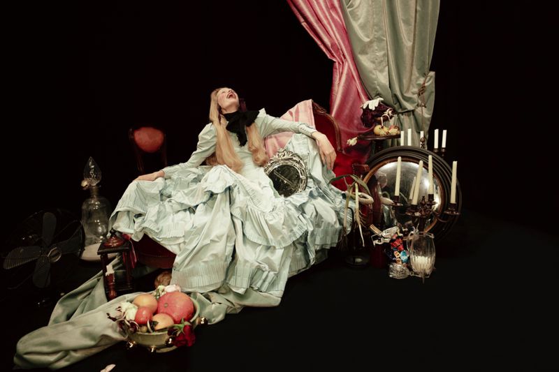 Woman in ruffled gown reclines amid props and drapery against a dark backdrop, creating a theatrical, vintage-inspired visual narrative.