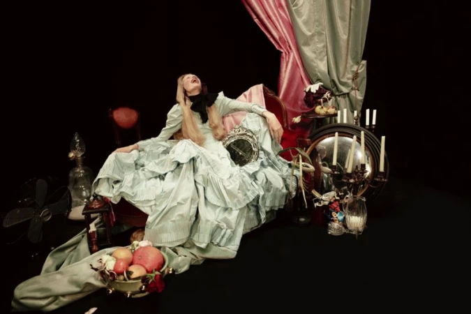 Woman in ruffled gown reclines amid props and drapery against a dark backdrop, creating a theatrical, vintage-inspired visual narrative.