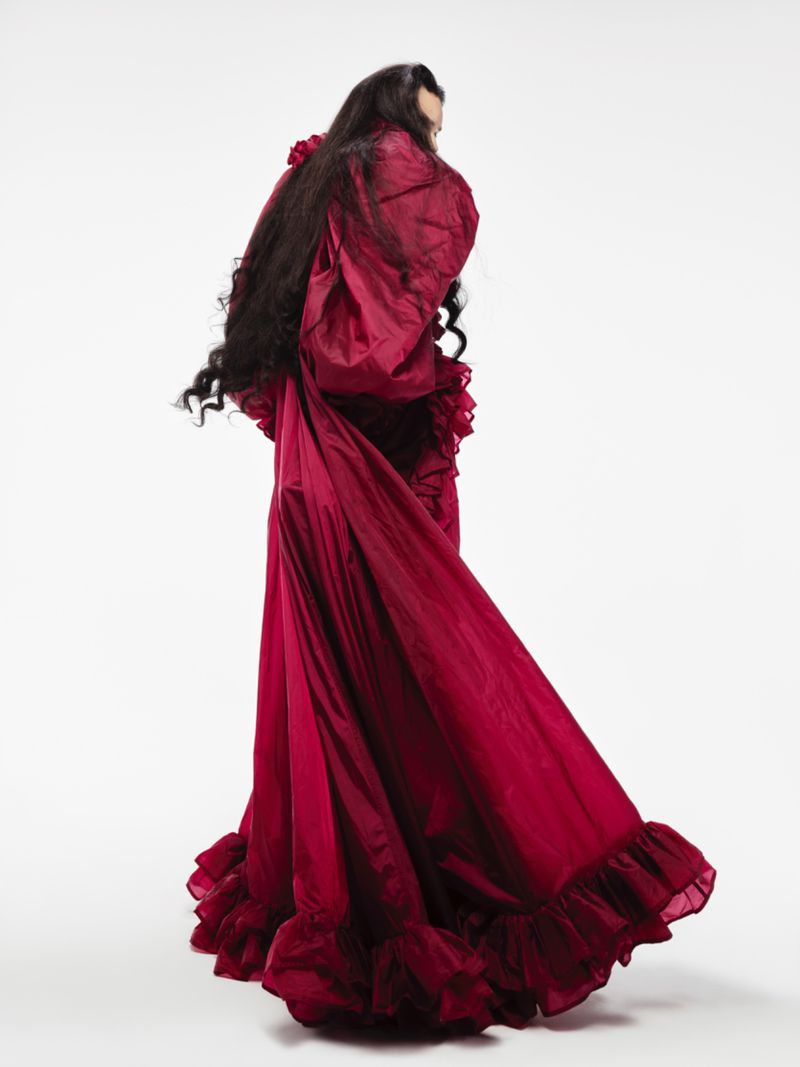 Woman with long dark hair poses in a flowing, ruffled, floor-length gown against a white backdrop in a fashion-style studio shot.