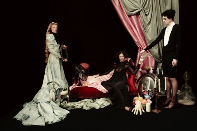 Three women in a stylized fashion shot. One woman is reclining on a red couch, another is standing and pouring a liquid, and one is holding a mirror.