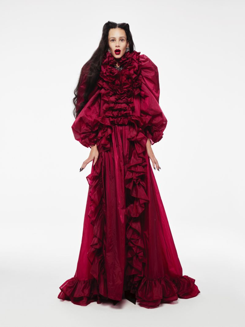 A person in a red, ruffled gown with a surprised expression, stands in a brightly lit studio with a stark white backdrop.
