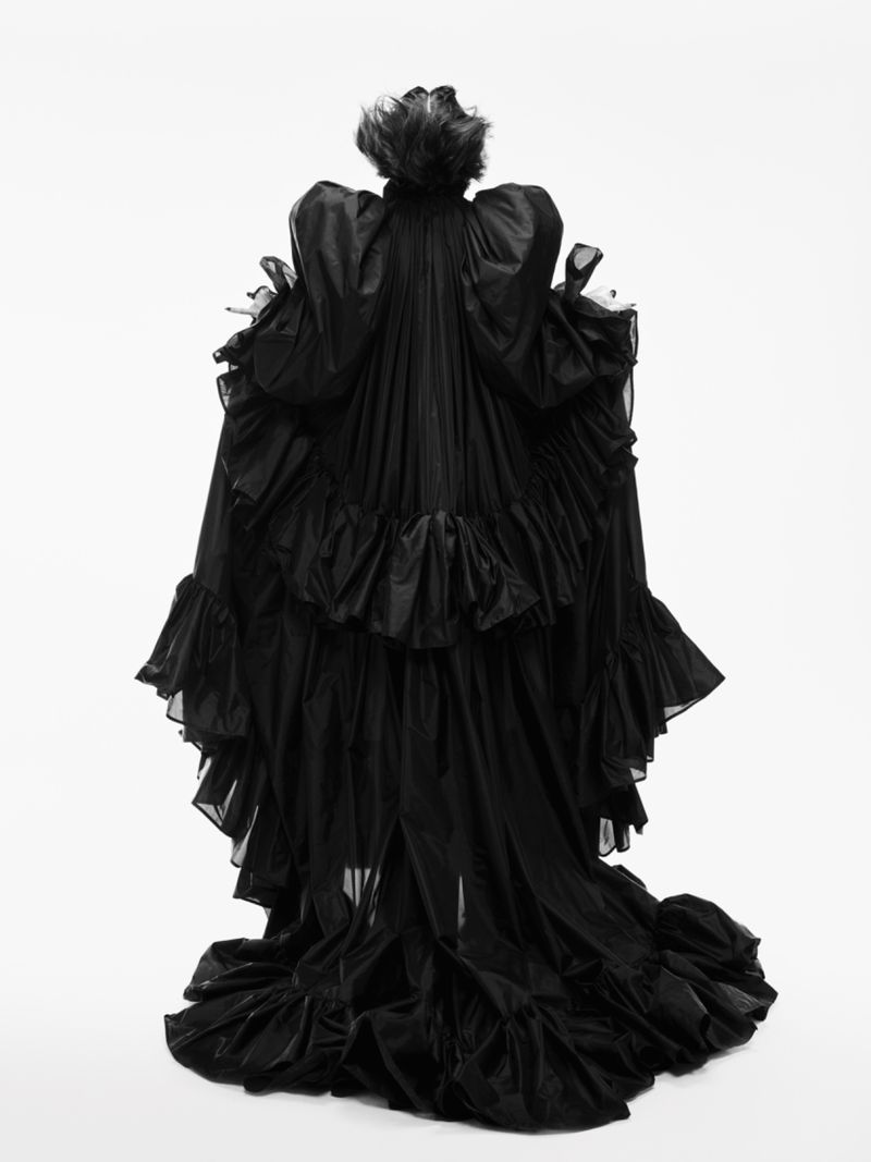 Back view of a person wearing a black, ruffled gown against a white backdrop in a studio setting. Dramatic fashion and photography.