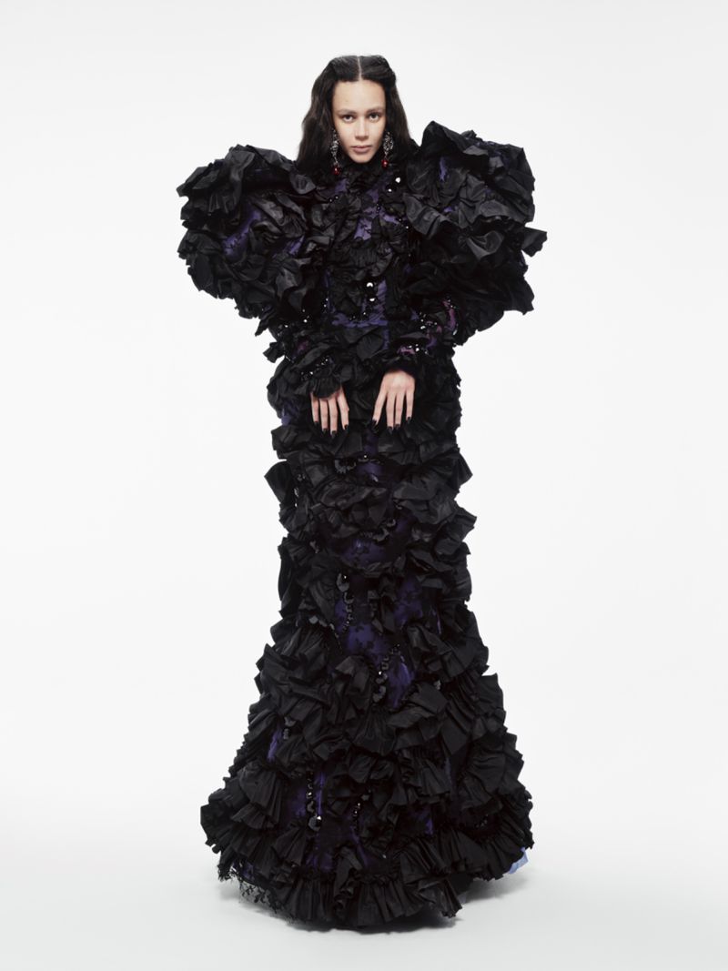 A model standing against a white background wearing an elaborate black ruffled gown with voluminous shoulders and deep purple accents.