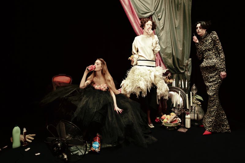 Three fashion models pose in a photo studio, two holding food and drink items, wearing elaborate dresses and dramatic makeup, with props.
