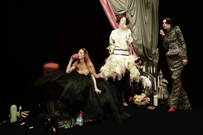 Three fashion models pose in a photo studio, two holding food and drink items, wearing elaborate dresses and dramatic makeup, with props.