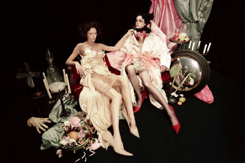 Two adult women in elegant, draped dresses pose together in a dark studio setting with draped fabric and antique props for a fashion editorial.