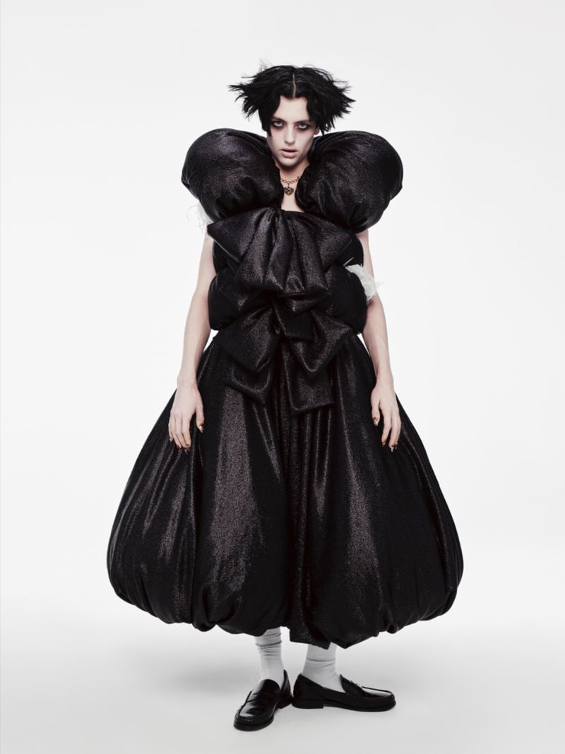 An adult female fashion model with dark hair poses in a dramatic black dress with a large bow against a white background in a studio shot.