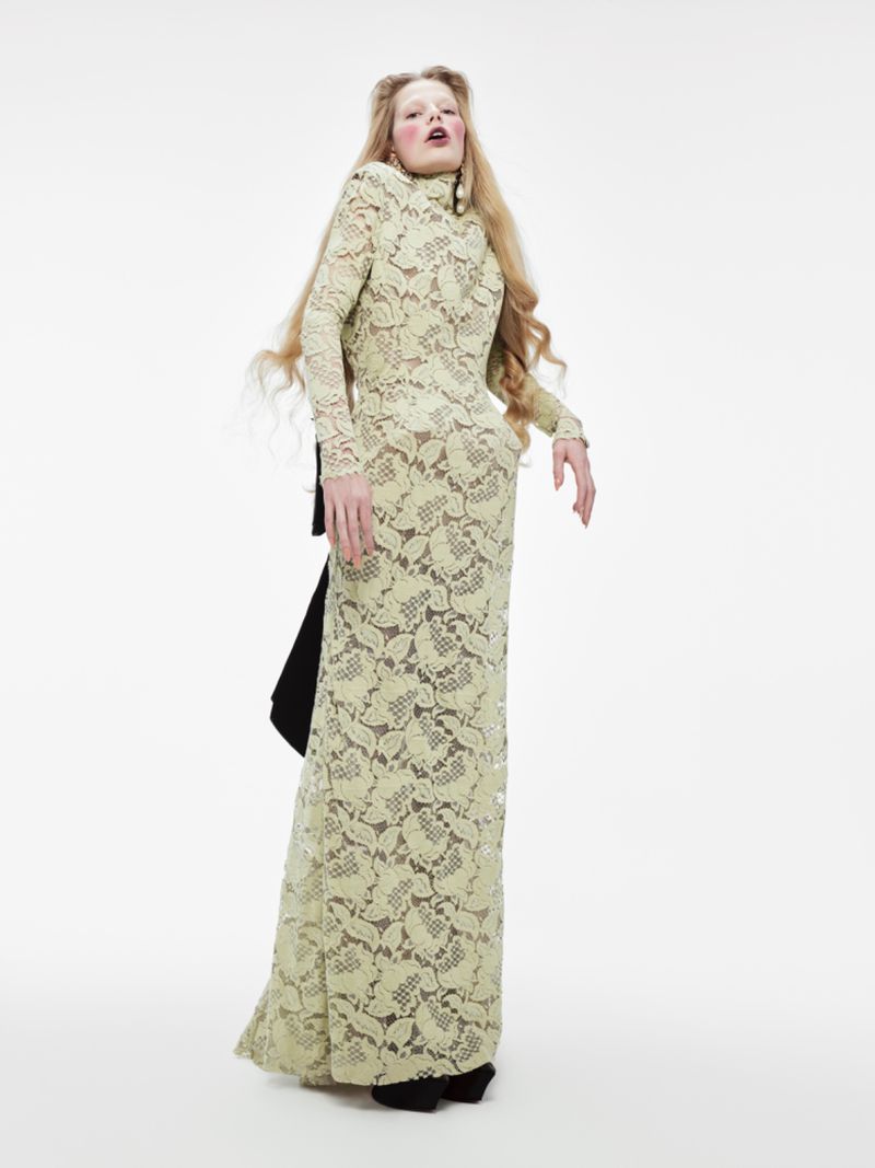 An adult female fashion model poses in a full-length pale yellow lace gown against a stark white backdrop in a full shot.