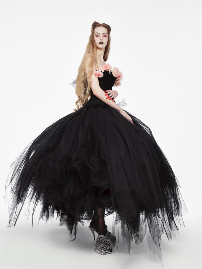 Blonde woman in a black tulle gown with roses on the bodice, posing in a bright studio with dramatic dark eye makeup and bold red nail polish.