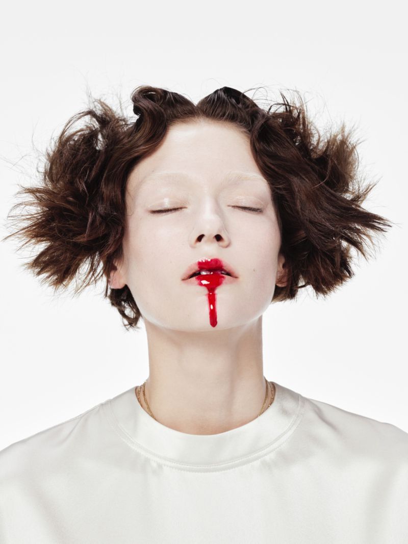 Close-up of a young woman with pale skin and short brown hair, eyes closed, red liquid dripping from her mouth, against a white background.