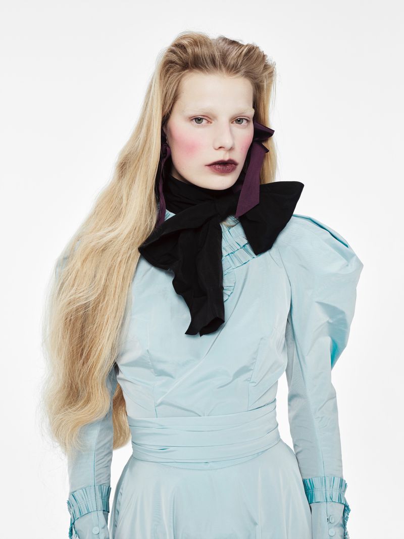 A woman with long blonde hair wears a light blue dress with a black bow, highlighted against a plain white backdrop in a fashion shot.