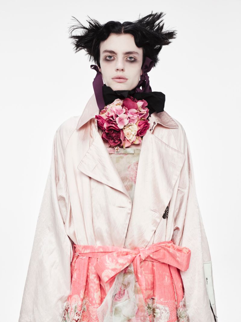 Androgynous person with pale complexion and dramatic makeup wearing a pale pink coat with floral accents in a white studio setting.