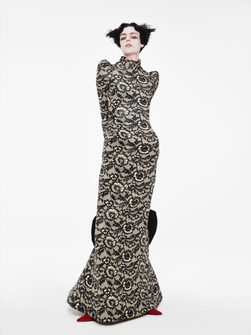 A slender woman in a floor-length black floral lace gown poses on a white seamless backdrop. She is wearing red pointed-toe heels.