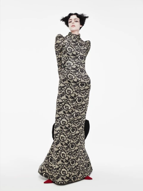 A slender woman in a floor-length black floral lace gown poses on a white seamless backdrop. She is wearing red pointed-toe heels.